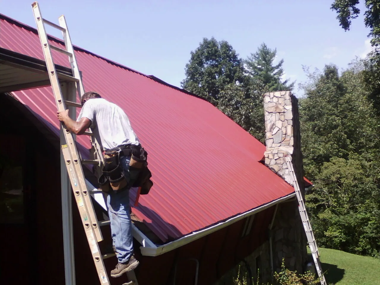 Roofer on a ladder beside a red metal roof and stone chimney — Soffit & Fascia Repair by Roofing Brothers in Jewell